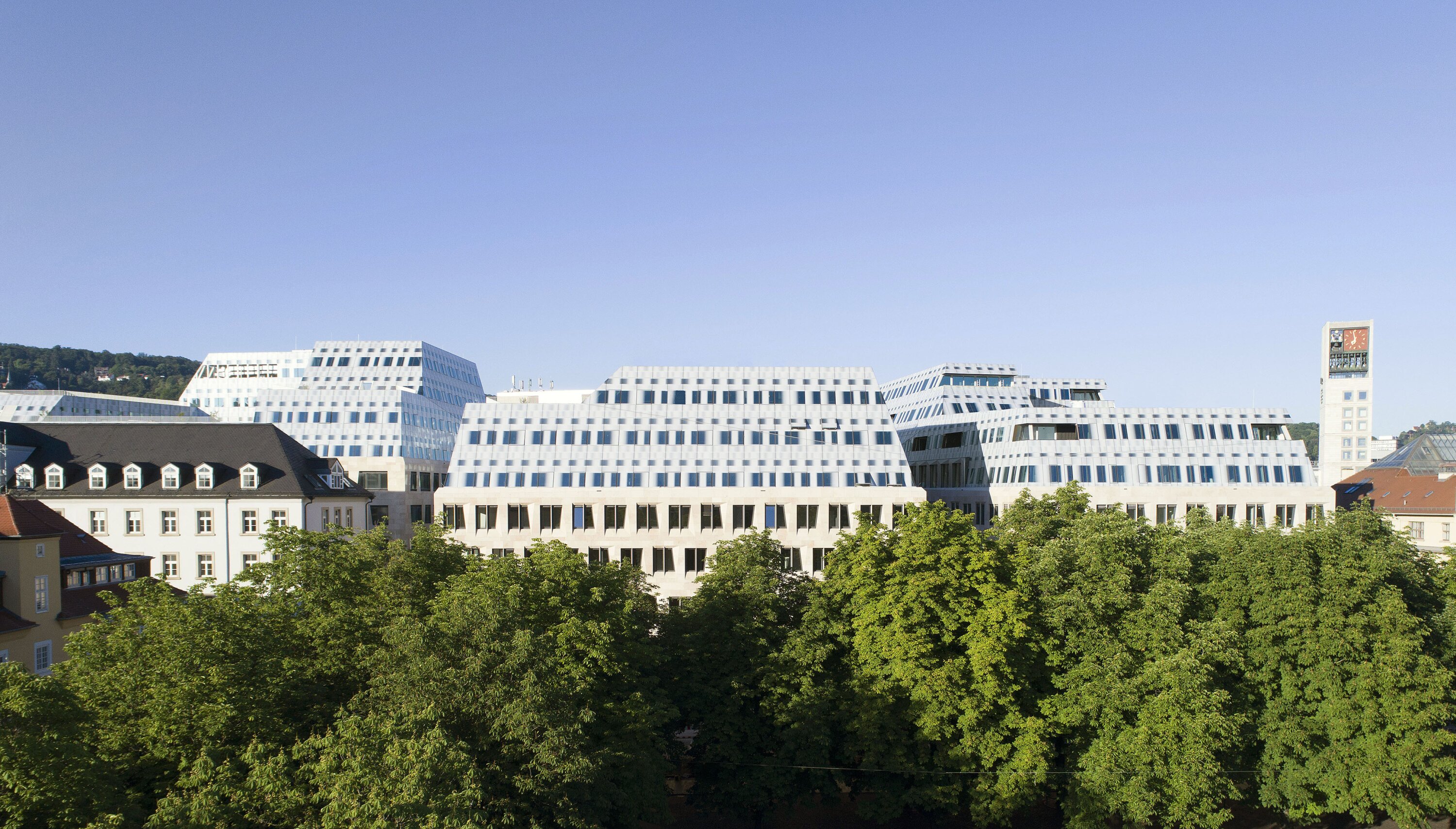 Project by Behnisch Architekturbuero, Dorotheen Quartier. Modern stepped office buildings behind a row of trees, with a clock tower and clear blue sky in the background.