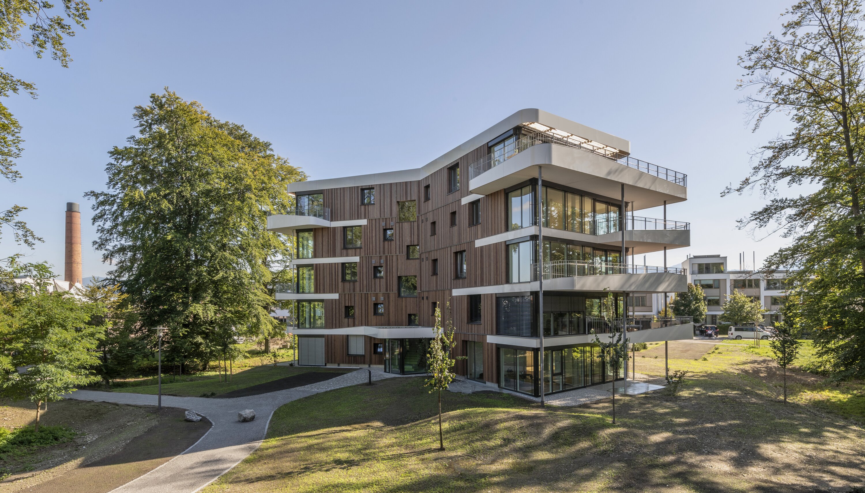 Project by Behnisch Architekturbuero, Y-Houses. A modern, multi-story residential building with a wooden facade and surrounding white balconies, set in a green park. A brick chimney is visible in the background.