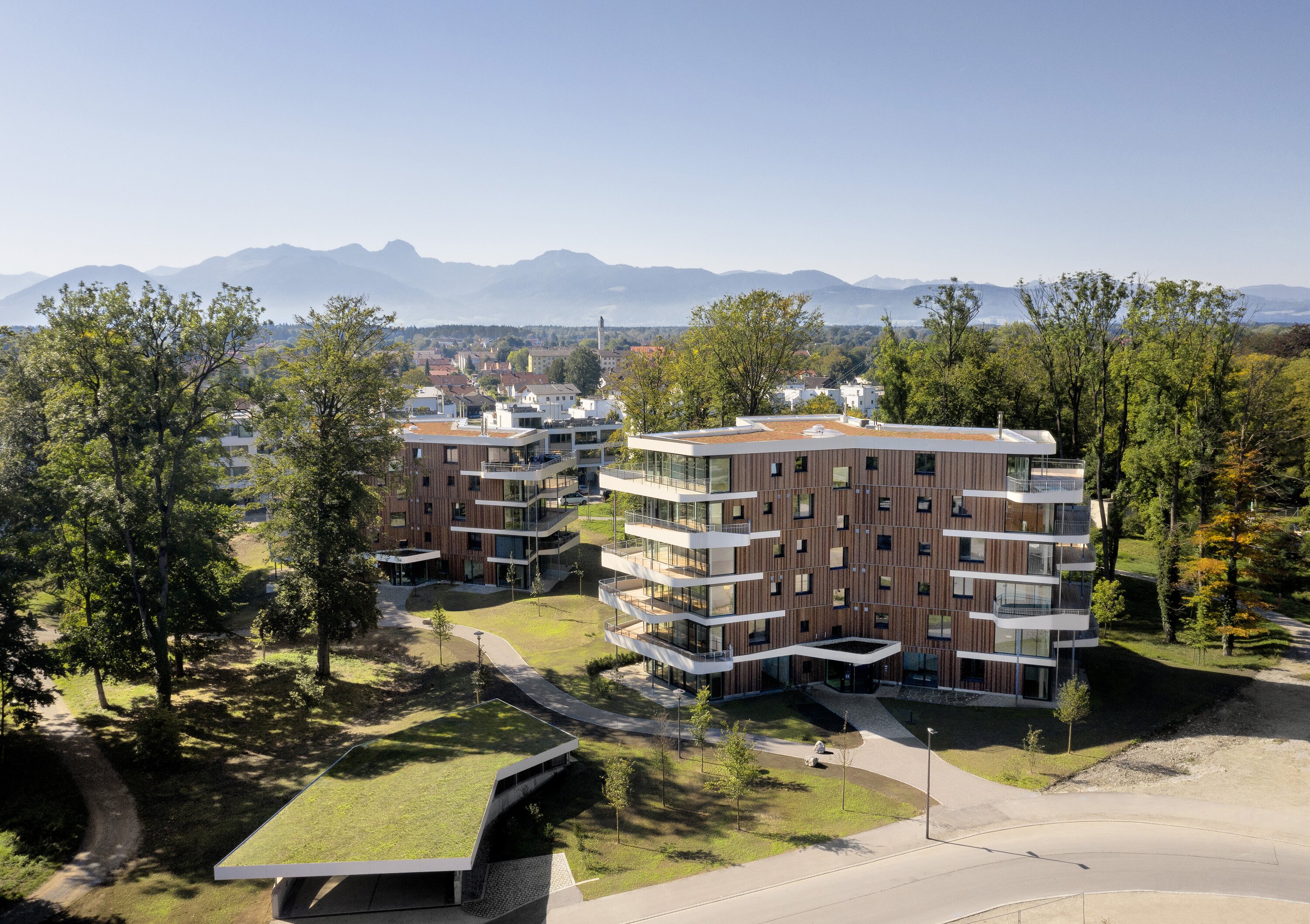 Project by Behnisch Architekturbuero, Y-Houses.Two modern apartment buildings with wooden facades and balconies, nestled in a well-maintained green area with a mountain panorama in the background.