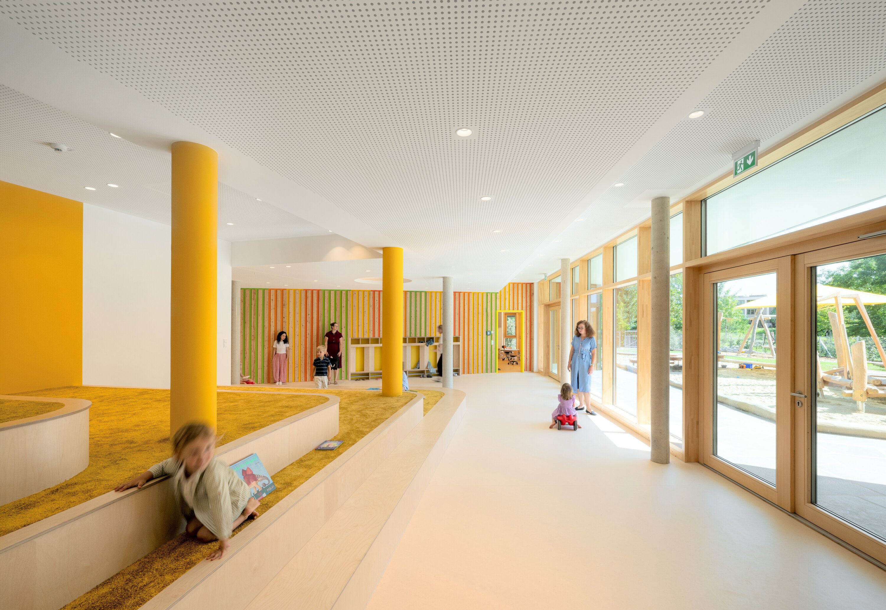 Project by Behnisch Architekturbuero, Conradty-Houses. Bright daycare interior with children and adults, curved carpeted play area and large windows.