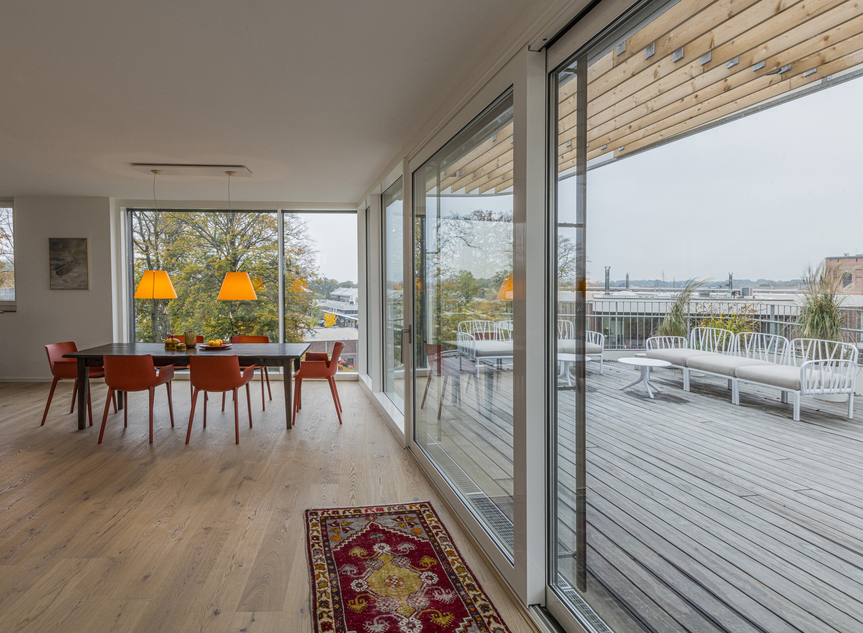 Project by Behnisch Architekturbuero, Y-Houses. Dining area with table and orange chairs. Generous floor-to-ceiling windows lead to a terrace with seating. An impression inside one of the penthouse apartments in the Y-buildings.