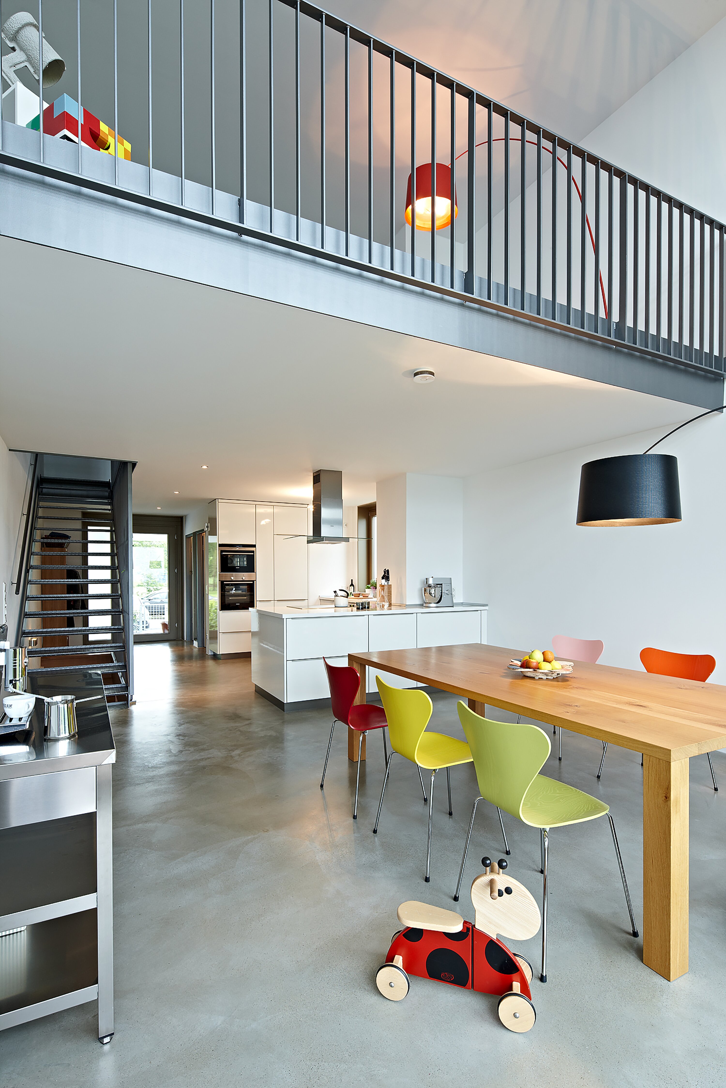 Project by Behnisch Architekturbuero, Loft Houses Kolbermoor. Modern open-plan kitchen and dining area with wooden table, colorful chairs, and mezzanine above. An impression into the interior of one of the Loft Houses.