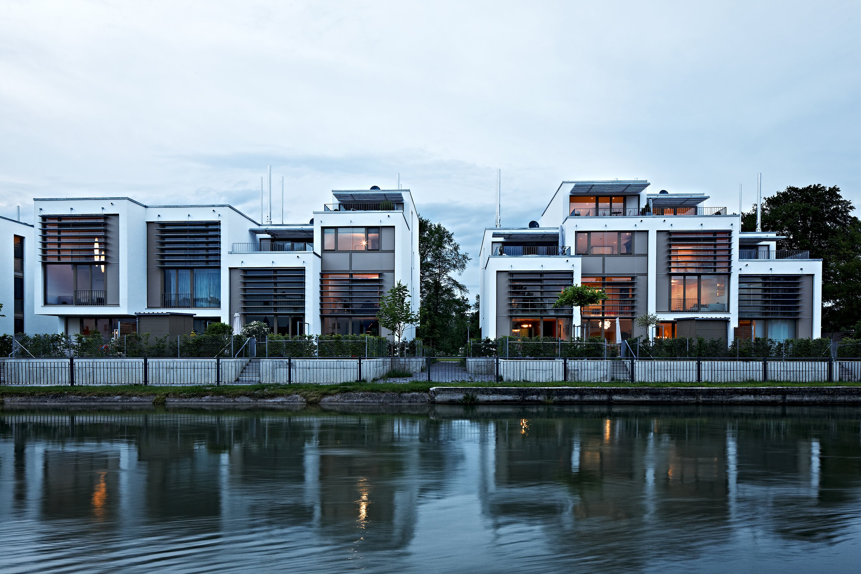 Project by Behnisch Architekturbuero, Loft Houses Kolbermoor. Front view of the modern white Loft Houses along the Mangfall Canal in an evening atmosphere.