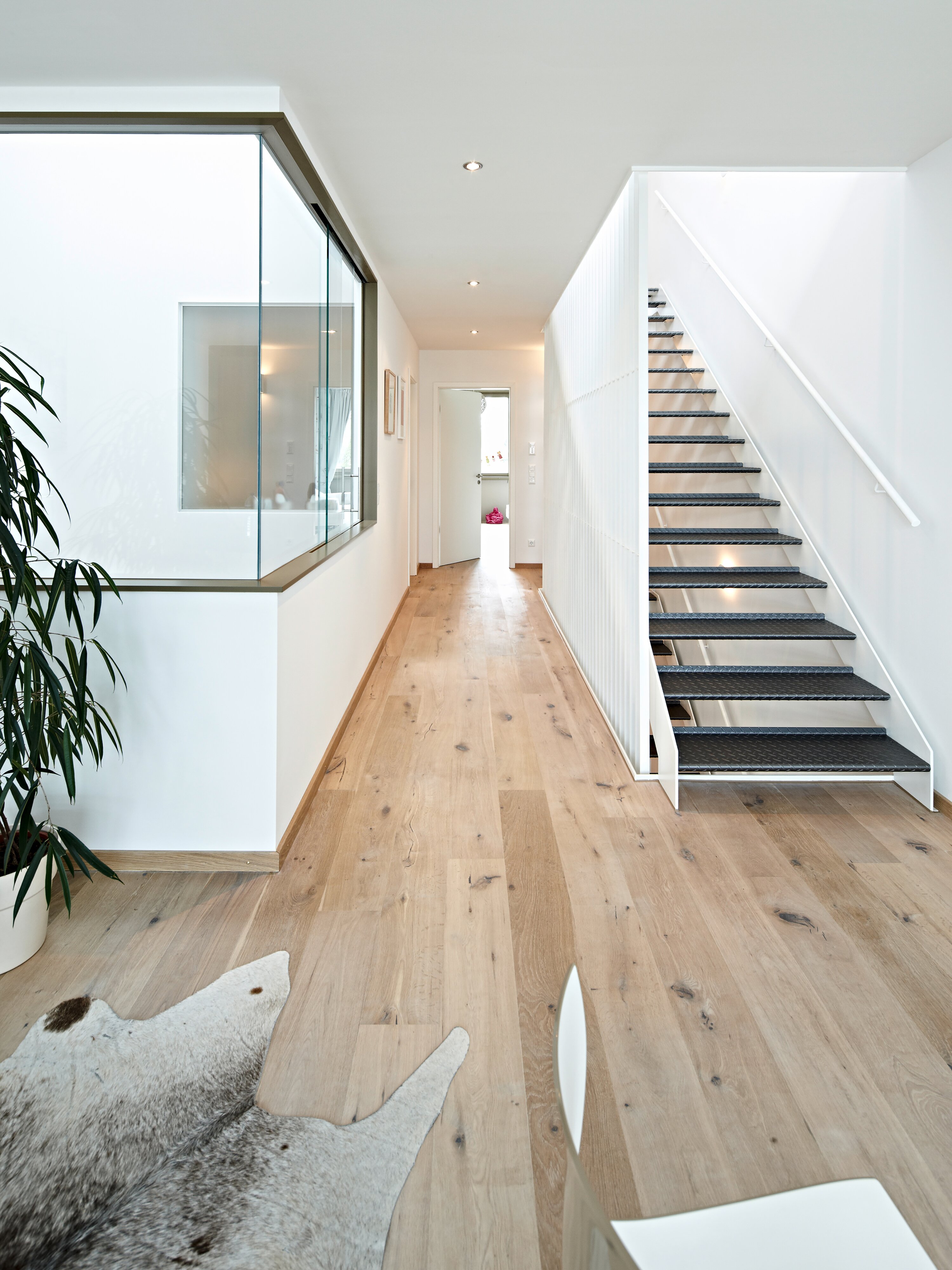 Project by Behnisch Architekturbuero, Loft Houses Kolbermoor. A bright, modern hallway with a wooden floor, a glass wall facing the courtyard, and an open staircase leading to the upper floor. An impression into the interior of one of the Loft Houses.