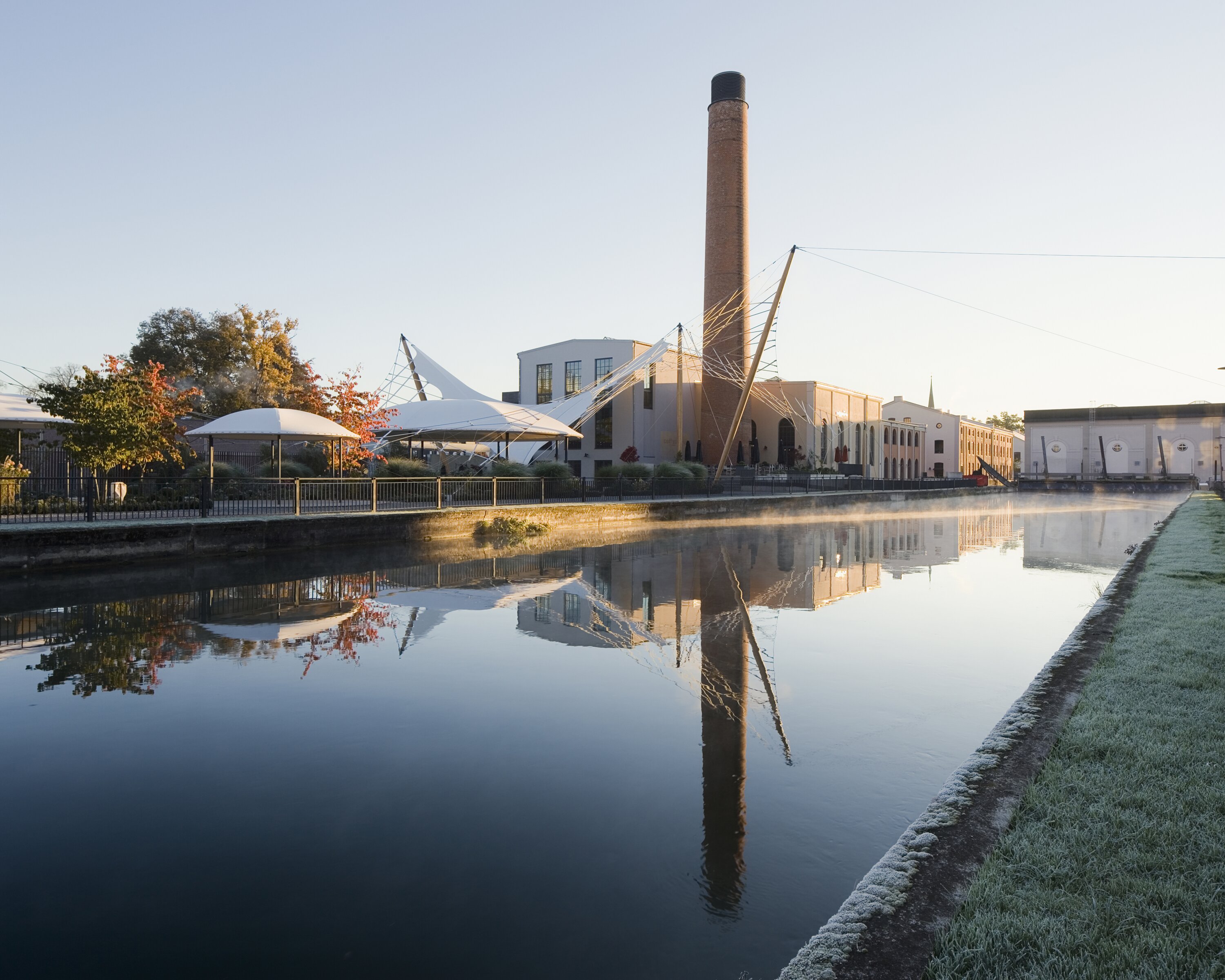Project by Behnisch Architekturbuero, QuestForum and Rosengarten. QuestForum and rose garden with a brick chimney in the background. Flexible event space covered by a rope net structure. Everything is reflected in the tranquil canal.
