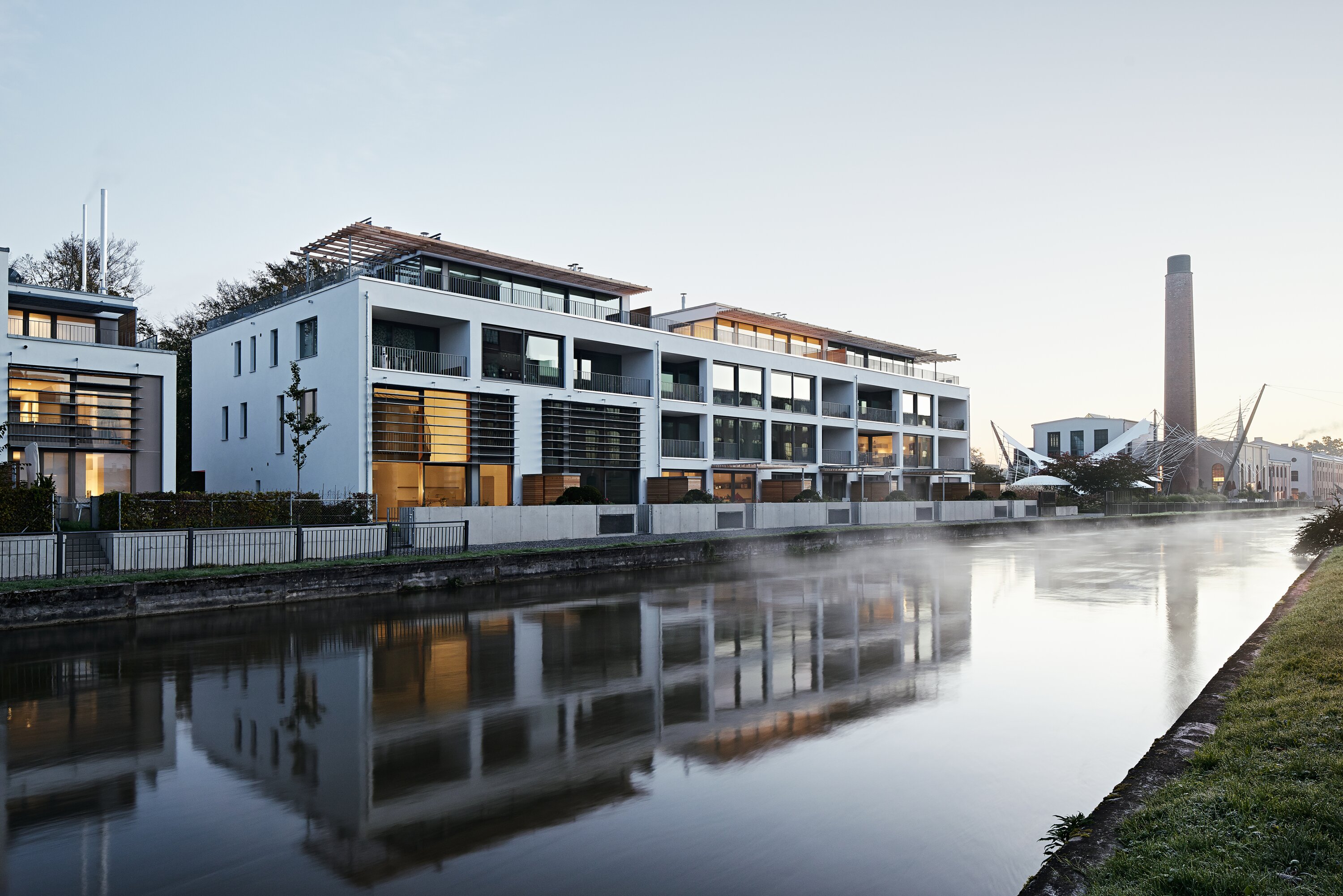 Project by Behnisch Architekturbuero, Apartment Building at the Rosengarten. Modern white apartment building with large windows and balconies reflected in a calm canal.