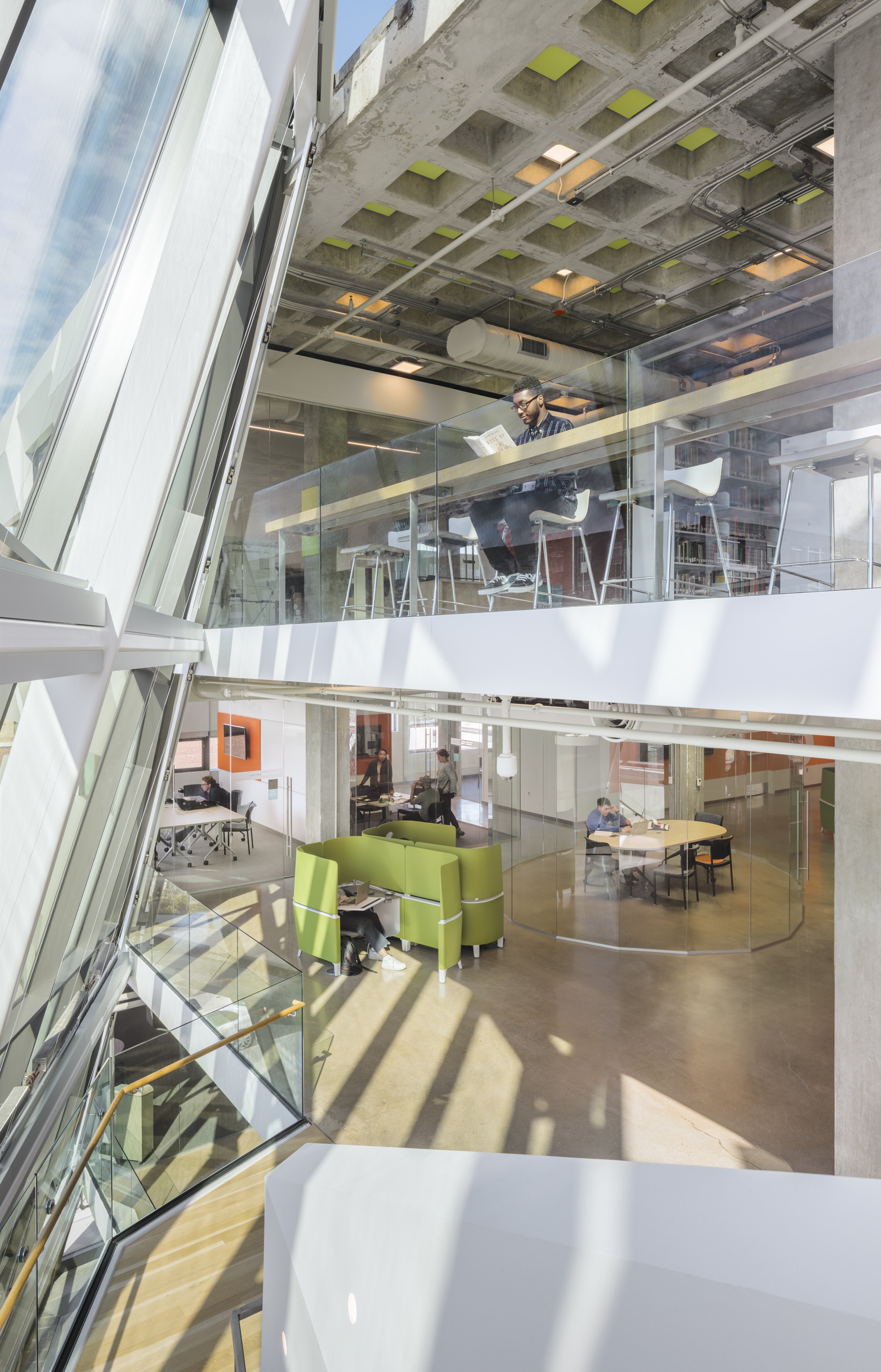 Project by Behnisch Architekturbuero, Robert Bogomolny Library. Multi-story atrium with a glass facade and exposed concrete ceiling. The employees work at tables, one of them on an upper mezzanine balcony.