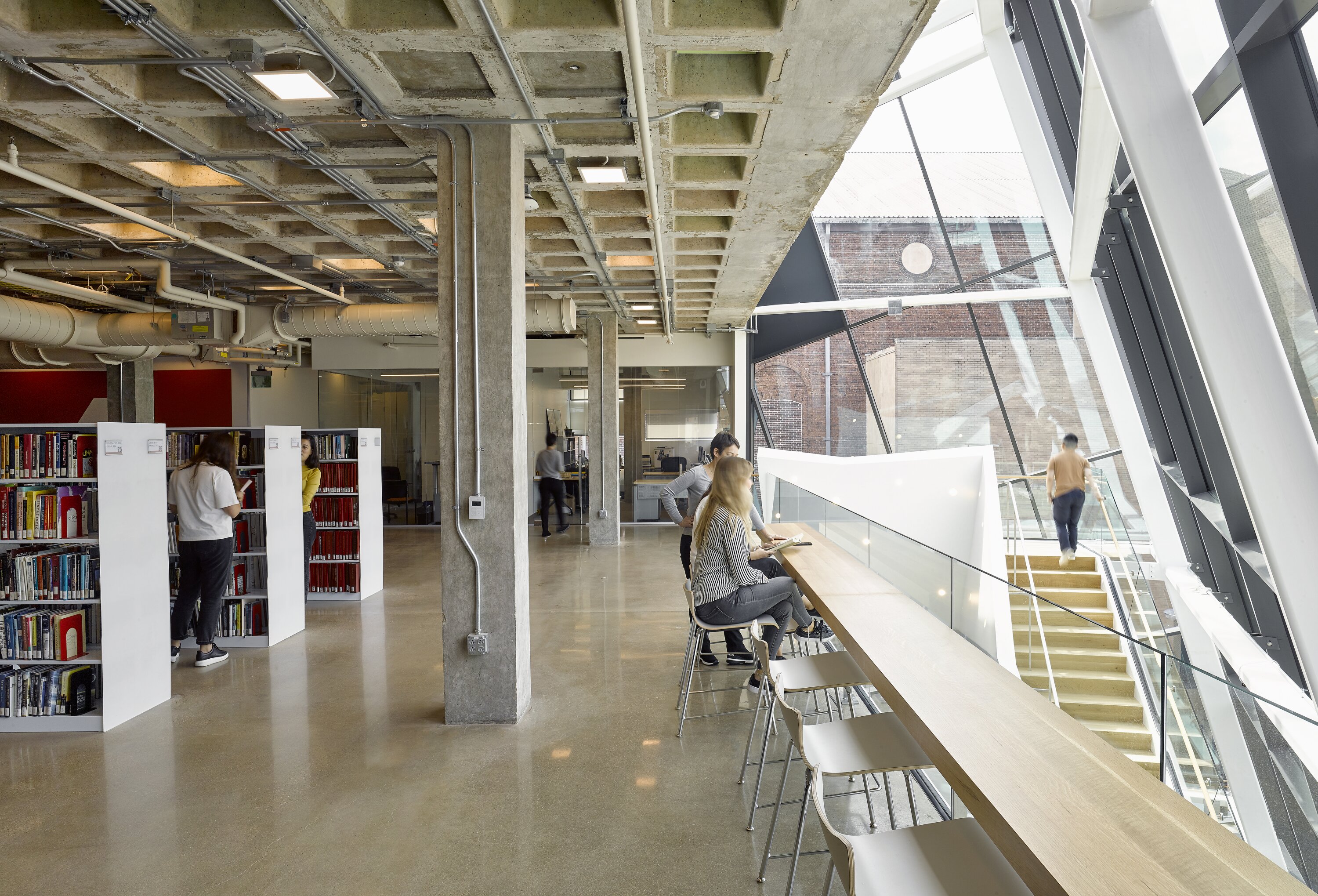 Project by Behnisch Architekturbuero, Robert Bogomolny Library. Library interior with bookshelves, exposed concrete ceiling, and window seating for reading. Behind it, a staircase and glass facade allow light to flood the room.