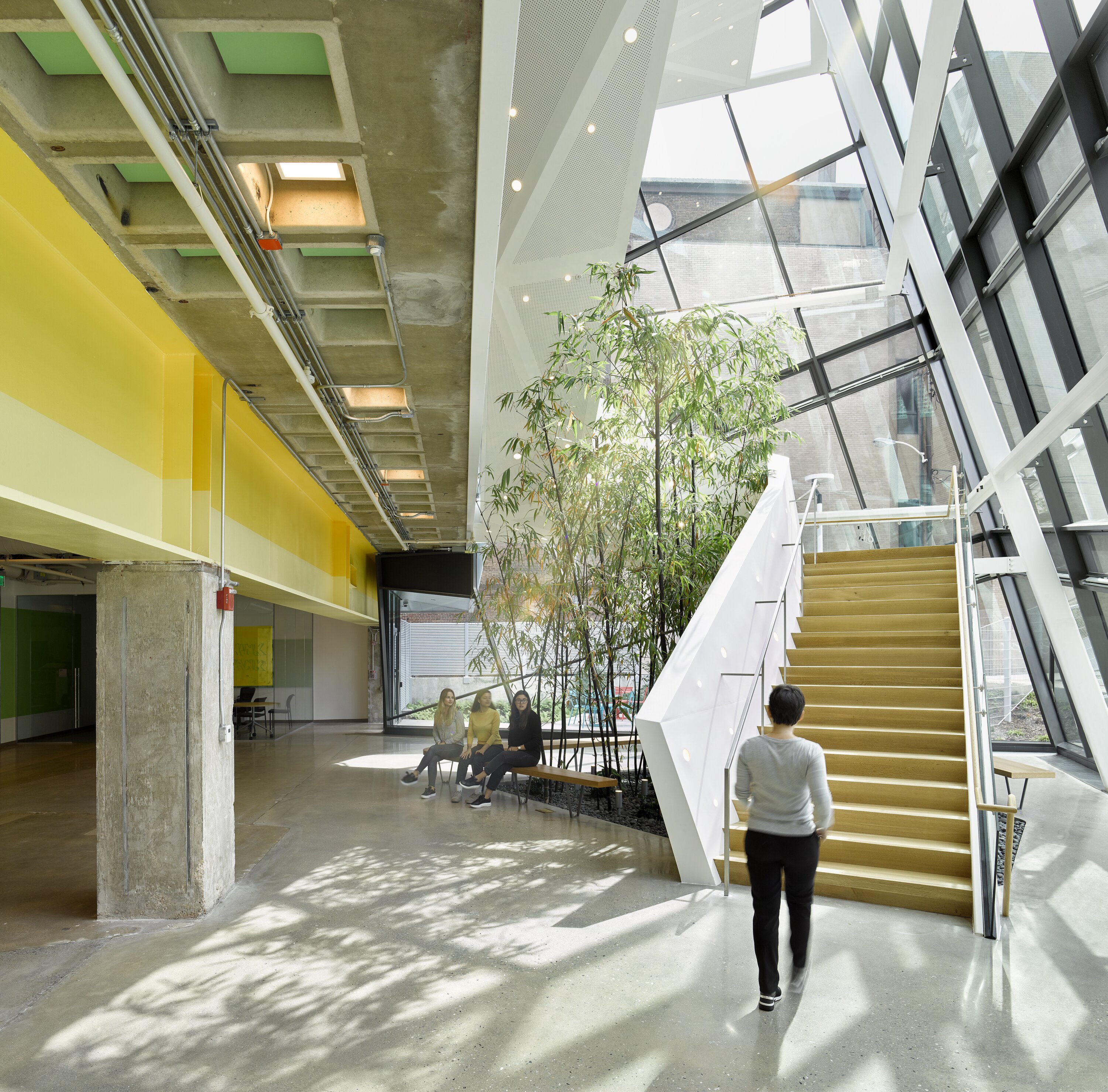 Project by Behnisch Architekturbuero, Robert Bogomolny Library. Bright atrium with a glass facade and staircase. Yellow beams and a grey exposed concrete ceiling. Houseplants, seating, and people sitting and walking around.